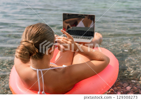 A woman is sitting on a red inflatable raft with a laptop on her lap. She is wearing a white bikini and sunglasses. Concept of relaxation and leisure. A woman is sitting on a red inflatable raft with a laptop on her lap. She is wearing a white bikini and sunglasses. Concept of relaxation and leisure. 129308273
