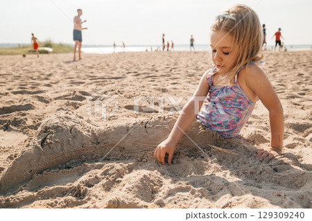 Young girl creates sandcastle at beach on sunny day while families enjoy nearby activities 129309240