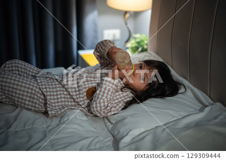 toddler baby girl drinking milk from bottle on a bed at night 129309444