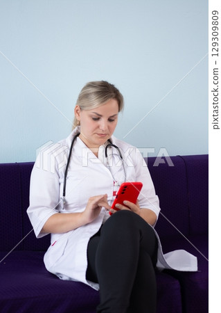 Female doctor in lab coat and stethoscope sitting with legs crossed and using a smartphone, focused and professional. Female doctor in lab coat and stethoscope sitting with legs crossed and using a smartphone, focused and professional. 129309809