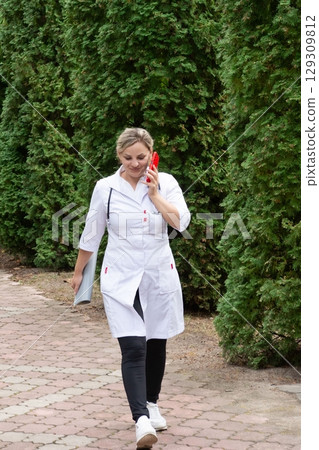 Medical professional in a lab coat walks along a brick path while talking on a cell phone surrounded by greenery on a sunny day. Medical professional in a lab coat walks along a brick path while talking on a cell phone surrounded by greenery on a sunny day. 129309812