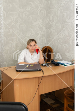 A blonde woman in a white coat is seated at a desk in a doctor's office, looking pensive while holding a red phone to her ear. A blonde woman in a white coat is seated at a desk in a doctor's office, looking pensive while holding a red phone to her ear. 129309813