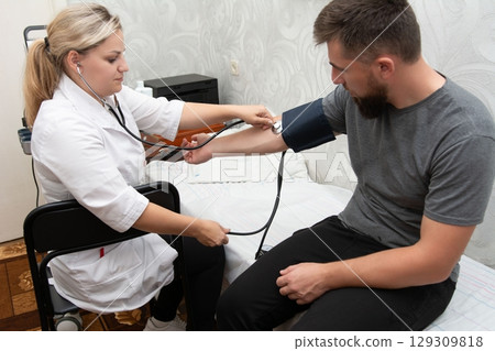 A woman in a white coat checks a man's blood pressure with a stethoscope and cuff on his arm. A monitor sits in the background. 129309818