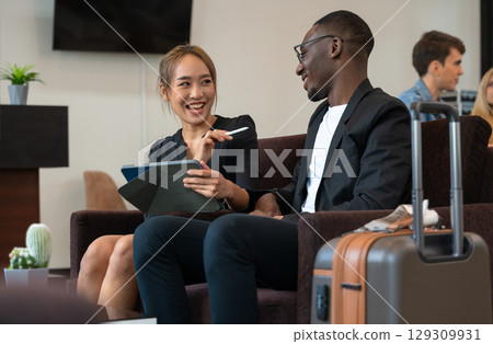 Cheerful two business travelers in airport lounge or hotel lobby. Asian woman using digital tablet PC while African man listens attentively Cheerful two business travelers in airport lounge or hotel lobby. Asian woman using digital tablet PC while African man listens attentively 129309931