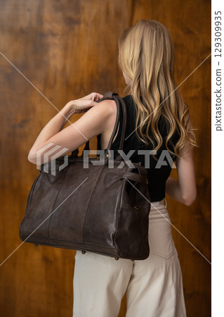 A beautiful fair-skinned model with wavy blonde hair in a half-body portrait, showcasing a minimalist black vest, wide-leg trousers, and a brown leather bag strap 129309935