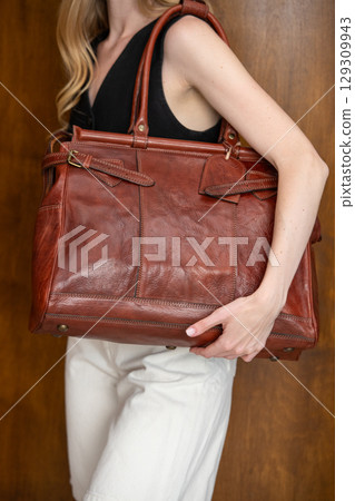 A beautiful fair-skinned model with wavy blonde hair in a half-body portrait, showcasing a minimalist black vest, wide-leg trousers, and a brown leather bag strap 129309943