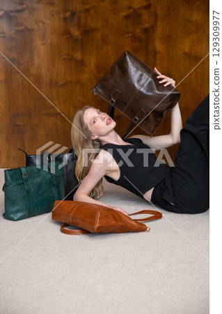 A beautiful fair-skinned model with wavy blonde hair in a dynamic full-body pose, lying on a beige carpet, showcasing multiple leather tote bags while holding one aloft. A beautiful fair-skinned model with wavy blonde hair in a dynamic full-body pose, lying on a beige carpet, showcasing multiple leather tote bags while holding one aloft. 129309977