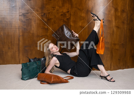 A beautiful fair-skinned model with wavy blonde hair in a dynamic full-body pose, lying on a beige carpet, showcasing multiple leather tote bags while holding one aloft. 129309988