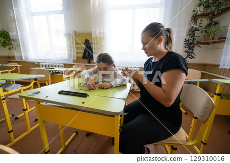 Teacher assisting student with learning activity at yellow desk in sunlit classroom, offering individualized educational attention. Teacher assisting student with learning activity at yellow desk in sunlit classroom, offering individualized educational attention. 129310016