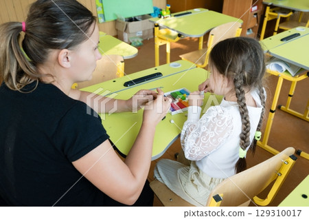 Teacher helping a student with colorful modeling clay at a light green school desk. The student has braids. Classroom environment. Teacher helping a student with colorful modeling clay at a light green school desk. The student has braids. Classroom environment. 129310017