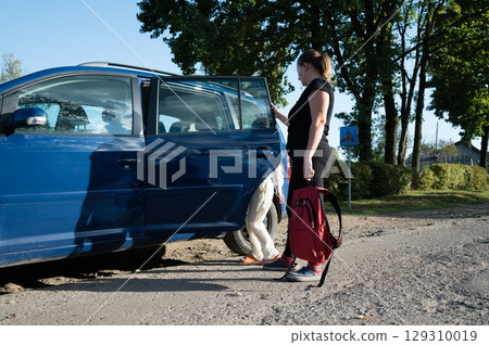 Woman opening a blue car door on a rural road, with another person exiting the vehicle near some trees on a sunny day. Backpack detail. Woman opening a blue car door on a rural road, with another person exiting the vehicle near some trees on a sunny day. Backpack detail. 129310019