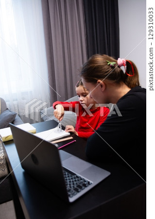 Child learning with tutor at home, using a drawing compass on paper, with a laptop in foreground, cozy, educational setting. Child learning with tutor at home, using a drawing compass on paper, with a laptop in foreground, cozy, educational setting. 129310033