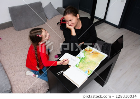 Focused young girl draws on a notepad as her mother lovingly watches, sitting together in a cozy spot for homework or creative pursuits. 129310034