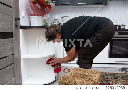 Woman cleaning an empty white kitchen corner cabinet with a cloth. Red roses bouquet on the kitchen counter. Home chores and cleaning. 129310089