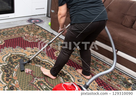Vacuuming a patterned rug barefoot in an apartment. Cleaning provides fresh living spaces and ensures a healthy indoor environment. 129310090