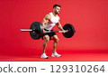 Muscular man in white tank top and black shorts lifting heavy barbell with focused expression on red background 129310264
