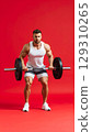 Muscular man in white tank top and shorts lifting heavy barbell with focus and strength on red background 129310265