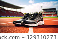 Pair of black and gray running shoes placed on red outdoor track with stadium background under bright sky 129310273