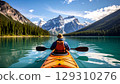Person paddling kayak on calm turquoise lake surrounded by pine forest and snow capped mountains under blue sky 129310276