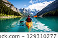 Person paddling kayak on turquoise lake surrounded by pine forest and snow capped mountains under blue sky 129310277