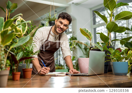 Young man sitting at the laptop in the flower shop Young man sitting at the laptop in the flower shop 129310336
