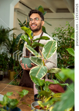Flower shop assistant presenting a plant in the pot and looking contented Flower shop assistant presenting a plant in the pot and looking contented 129310426