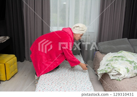 Woman making a daybed in a cozy bedroom. She is wearing a towel around her head and a red robe, creating a comfortable domestic scene. Woman making a daybed in a cozy bedroom. She is wearing a towel around her head and a red robe, creating a comfortable domestic scene. 129310431
