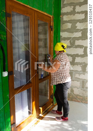 Cleaning the window glass of the wooden framed windows. Woman wearing headscarf, using cloth and cleaner for spotless shine. Door, green walls. Cleaning the window glass of the wooden framed windows. Woman wearing headscarf, using cloth and cleaner for spotless shine. Door, green walls. 129310437