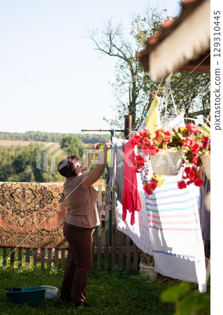 Woman hanging laundry on an outdoor clothesline. Fresh clothes drying under the sun. A sunny day chore in a grassy backyard. Woman hanging laundry on an outdoor clothesline. Fresh clothes drying under the sun. A sunny day chore in a grassy backyard. 129310445