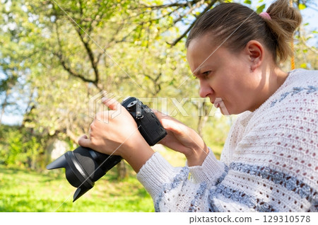 Focused photographer reviewing images on a digital camera screen outdoors. Woman, equipment, and nature backdrop. Capturing moments. 129310578