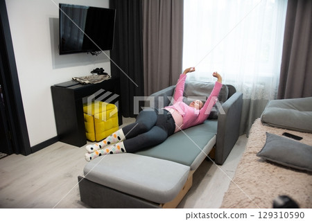 Relaxing at Home: Woman stretches out on a sofa bed in a cozy room, enjoying a moment of rest and rejuvenation indoors. 129310590