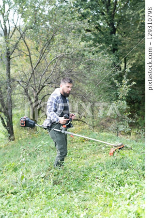 Man using a string trimmer to cut tall grass and weeds on a residential lawn in the backyard with trees, and he is wearing working clothes. Man using a string trimmer to cut tall grass and weeds on a residential lawn in the backyard with trees, and he is wearing working clothes. 129310798