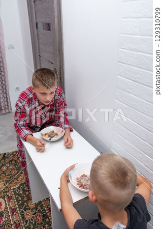 Two young boys are seen indoors sharing a meal, likely breakfast, enjoying a bonding moment at home in a brightly lit space. 129310799