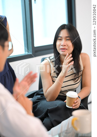Asian employee sitting in an office, holding a coffee cup and appearing to gossip with a colleague Asian employee sitting in an office, holding a coffee cup and appearing to gossip with a colleague 129310842
