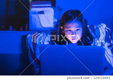 Asian girl sitting on the couch watching laptop at night. Parental control, screen addiction, and bedtime behavior. Asian girl sitting on the couch watching laptop at night. Parental control, screen addiction, and bedtime behavior. 129310991