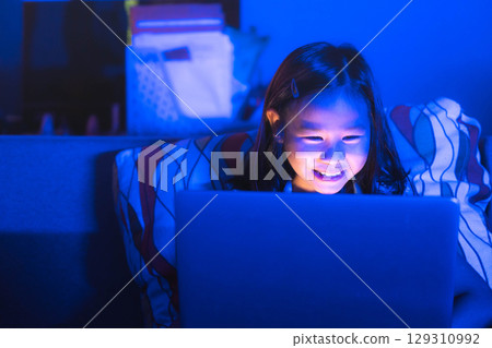 Asian girl sitting on the couch watching laptop at night. Parental control, screen addiction, and bedtime behavior. 129310992