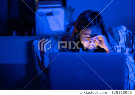 Asian girl sitting on the couch watching laptop at night. Parental control, screen addiction, and bedtime behavior. Asian girl sitting on the couch watching laptop at night. Parental control, screen addiction, and bedtime behavior. 129310996