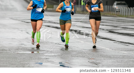 three young female athletes running marathon in cold weather on wet road three young female athletes running marathon in cold weather on wet road 129311348