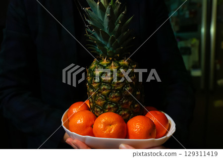 A festive New Year's night scene with hands holding a fresh pineapple and vibrant tangerines. The image symbolizes new beginnings and good luck for the new year A festive New Year's night scene with hands holding a fresh pineapple and vibrant tangerines. The image symbolizes new beginnings and good luck for the new year 129311419