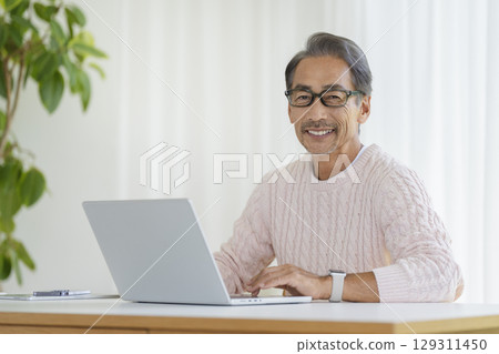 一位 60 多歲的男子正在使用筆記型電腦，面帶微笑，看著鏡頭 129311450