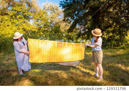 Caucasian mom and her young daughter unfold yellow picnic mat in green city park on sunny day. Family weekend, outdoor fun and casual lifestyle. 129311706