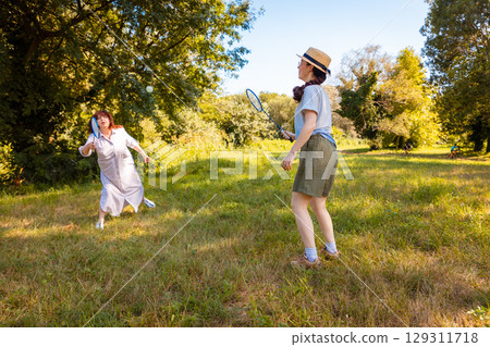 Happy young girl and her mother enjoying badminton in the green park during vacation. Active lifestyle, healthy family recreation and fun sport activity in nature. Wide shot 129311718
