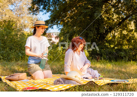 Mature woman and her young daughter spending warm afternoon on a plaid in nature. Concept of family holidays and mental health care. Mature woman and her young daughter spending warm afternoon on a plaid in nature. Concept of family holidays and mental health care. 129311720