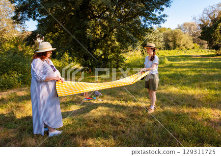 Middle aged mother and teenage Caucasian daughter spread picnic blanket on grass in summer park. Family bonding, vacation time and outdoor leisure concept. Middle aged mother and teenage Caucasian daughter spread picnic blanket on grass in summer park. Family bonding, vacation time and outdoor leisure concept. 129311725