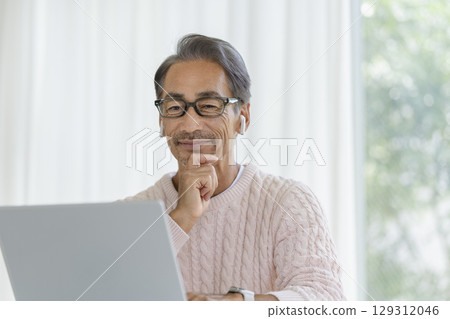 A man in his 60s wearing earphones looking at a laptop screen A man in his 60s wearing earphones looking at a laptop screen 129312046