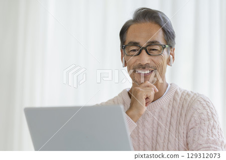 一位 60 多歲的男子戴著耳機看著筆記型電腦螢幕 129312073