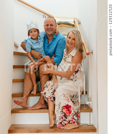 A cheerful family poses together on a staircase with a gold number 5 balloon, celebrating their son's fifth birthday in a cozy home setting 129312520