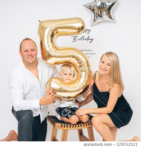 Smiling parents and their young son celebrate his 5th birthday in a bright studio with festive balloons. Ideal for family lifestyle, birthday greeting cards, and parenting content. Smiling parents and their young son celebrate his 5th birthday in a bright studio with festive balloons. Ideal for family lifestyle, birthday greeting cards, and parenting content. 129312521