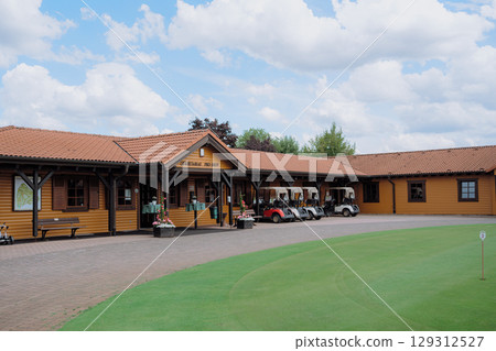 Golf pro shop building with several golf carts parked outside, adjacent to a putting green under a bright sky. The image captures a serene golf course environment. Golf pro shop building with several golf carts parked outside, adjacent to a putting green under a bright sky. The image captures a serene golf course environment. 129312527