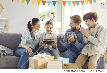 Happy girl in party hat celebrating birthday with family at home, looking at cake with candles 129312578